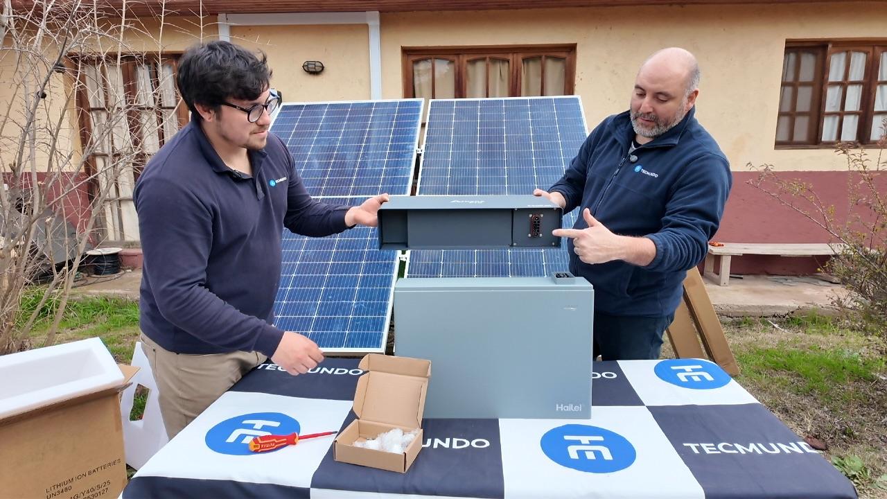 Dos técnicos de Tecmundo revisan una batería sobre una mesa, con paneles solares detrás